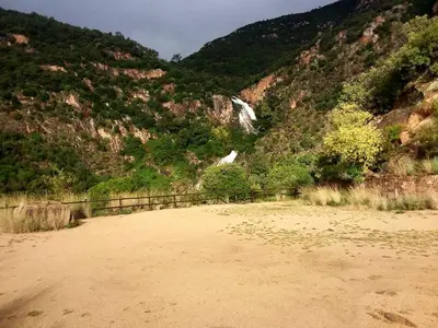 a dirt field in front of a mountain with a waterfall at Càmping Parc Gualba in gualba de Dalt